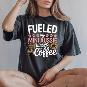 May include: Dark gray t-shirt with the text "FUELED by MINI AUSSIE kisses and coffee" in white, pink, and gold. The design includes paw prints, a coffee cup, and a coffee cup graphic.