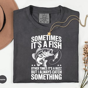 May include: Graphite-colored t-shirt with the text "SOMETIMES IT'S A FISH, OTHER TIMES IT'S A BUZZ BUT I ALWAYS CATCH SOMETHING" and a fish graphic. The shirt has a crew neck and short sleeves. A gold necklace with a star pendant is on the shirt.