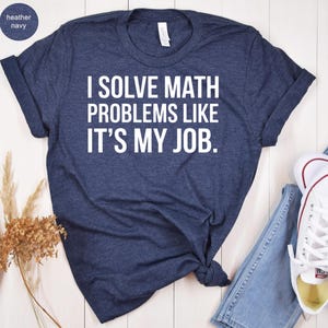 May include: Heather navy blue t-shirt with the text "I SOLVE MATH PROBLEMS LIKE IT'S MY JOB" in white. The shirt is styled with rolled-up sleeves and a knot at the bottom. A pair of jeans and white sneakers are visible.