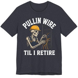May include: Dark gray t-shirt with the text "PULLIN WIRE TIL I RETIRE". The graphic features a skeleton wearing a yellow hard hat and overalls, working with wires in an electrical panel.