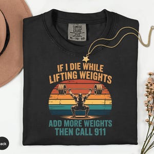 May include: Black t-shirt with a graphic of a weightlifter lifting weights against a sunset backdrop. The text reads "IF I DIE WHILE LIFTING WEIGHTS ADD MORE WEIGHTS THEN CALL 911". A gold necklace with a star pendant is draped on the shirt.