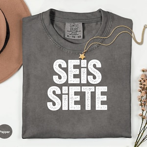 May include: A dark gray t-shirt with the words "SEIS SIETE" in large white letters. A gold necklace with a star pendant rests on the shirt. The shirt is labeled XL-TG-EG.