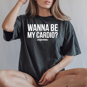 May include: Dark gray t-shirt with the text "WANNA BE MY CARDIO? (respectfully)" in white block letters. The shirt has short sleeves and a relaxed fit. The shirt is a casual wear item.