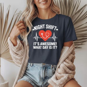 May include: Navy blue t-shirt with white text that reads "NIGHT SHIFT" above a red heart with a stethoscope and a heartbeat line. Below the heart, the text reads "IT'S AWESOME! WHAT DAY IS IT?"