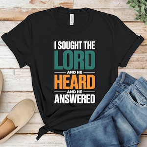 May include: Black t-shirt with the text "I Sought The Lord And He Heard And He Answered" in white, teal, and orange. The shirt is paired with blue jeans and beige slip-on shoes.