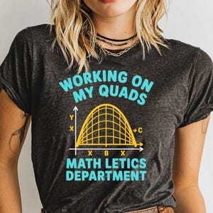 May include: Dark gray t-shirt with the text "WORKING ON MY QUADS" in turquoise. Below is a yellow graph with grid lines and the words "MATH LETICS DEPARTMENT" in turquoise. The shirt is a crew neck.