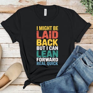 May include: Black t-shirt with the text "I MIGHT BE LAID BACK BUT I CAN LEAN FORWARD REAL QUICK" in red, yellow, teal, and white. The shirt is paired with blue jeans and beige slip-on shoes.