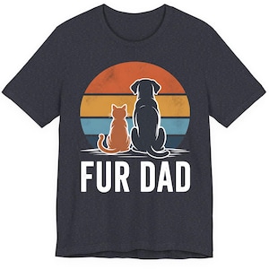 May include: Dark gray t-shirt featuring a graphic of a cat and dog silhouette sitting together in front of a sunset. The text "FUR DAD" is printed in white below the graphic. The shirt is made of a soft material.