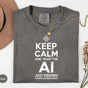 May include: A gray Comfort Colors t-shirt with the text "KEEP CALM AND TRUST THE AI JUST KIDDING ALWAYS DOUBLE-CHECK" in white. A gold necklace with a star pendant rests on the shirt. The shirt is size XL-TG-EG.