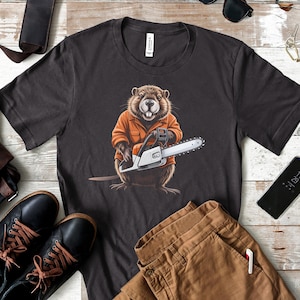 May include: Dark gray t-shirt featuring a cartoon beaver wearing an orange work shirt and holding a chainsaw. The beaver has a friendly expression. The shirt is a crew neck and is made of a soft material.