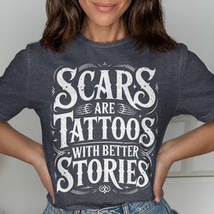 May include: Dark gray t-shirt with the quote "Scars are Tattoos with Better Stories" in white, stylized lettering. The design features decorative flourishes and a vintage aesthetic. The shirt is a crew neck.
