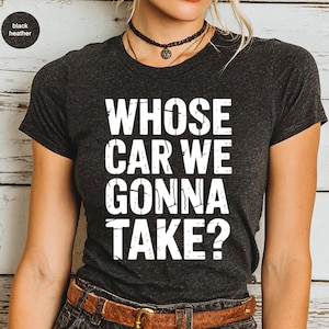 May include: Dark heather gray t-shirt with the text "WHOSE CAR WE GONNA TAKE?" in large white letters. The shirt has a crew neck and short sleeves. The model is wearing a brown belt and a black choker necklace.