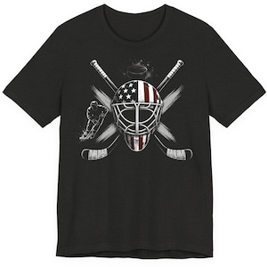 May include: Dark gray t-shirt featuring a hockey-themed graphic. The design includes a goalie mask with a US flag design, crossed hockey sticks, a hockey puck, and a hockey player silhouette. The graphic is in shades of white, gray, and red.