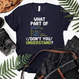 May include: A navy blue t-shirt with white and green pixelated text that reads "WHAT PART OF / DON'T YOU / UNDERSTAND?". The shirt features code snippets. The shirt is displayed with black pants and a leather bag.
