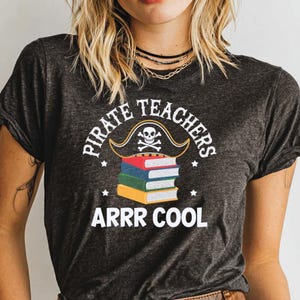 May include: Dark gray t-shirt with the text "PIRATE TEACHERS ARRR COOL". The design features a pirate hat with a skull and crossbones above a stack of colorful books. The shirt is a crew neck with rolled-up sleeves.