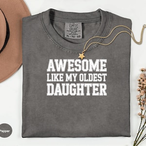May include: A gray t-shirt with the text "AWESOME LIKE MY OLDEST DAUGHTER" in white block letters. A gold necklace with a star pendant is draped across the shirt. The shirt is a Comfort Colors brand, size XL-TG-EG.