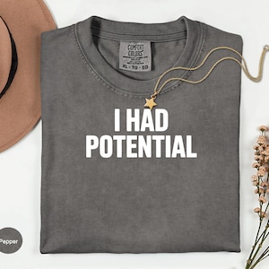 May include: Pepper-colored t-shirt with the text "I HAD POTENTIAL" in white block letters. A gold necklace with a star pendant rests on the shirt. A brown hat and sprigs of pink flowers are in the background.