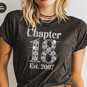 May include: Black heather t-shirt with the text "Chapter 18 Est. 2007" in a sparkling, rhinestone-like font. The numbers are embellished with a crystal-like design. The shirt is a dark gray color.