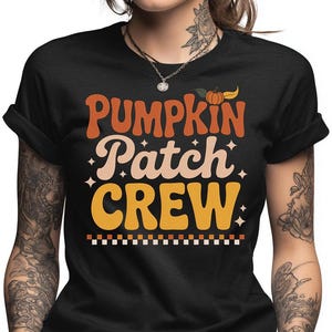 May include: Black t-shirt with the words "PUMPKIN Patch CREW" in orange, cream, and yellow lettering. A small pumpkin graphic is above the word "CREW". The shirt has a retro, fall-themed design.