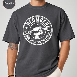 May include: Graphite-colored t-shirt with a white circular graphic. The graphic features the word "PLUMBER" above a hand holding a wrench, with the text "THE MAN THE MYTH THE LEGEND" around the edge. The shirt has a relaxed fit.