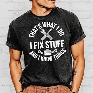 May include: Dark gray t-shirt with the text "THAT'S WHAT I DO, I FIX STUFF AND I KNOW THINGS" in a white, circular design. The design includes a wrench, screwdriver, toolbox, and gears.