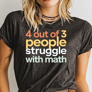 May include: Dark gray t-shirt with the text "4 out of 3 people struggle with math" in orange, yellow, and white. The shirt is a crew neck and has rolled-up sleeves.