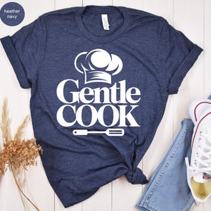 May include: Heather navy blue t-shirt with the words "Gentle Cook" in white, along with a chef's hat and spatula graphic. The shirt is folded at the bottom and paired with blue jeans and white sneakers.