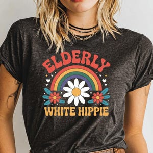 May include: Dark gray t-shirt with a retro design. The design features the word "ELDERLY" in red, arched over a rainbow, a white daisy, and the words "WHITE HIPPIE" in yellow.
