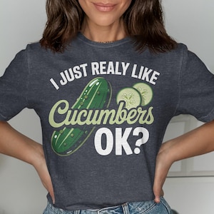 May include: Dark gray t-shirt with the text "I JUST REALLY LIKE Cucumbers OK?" in white and green. The graphic features a cucumber and cucumber slices. The shirt is a casual style.