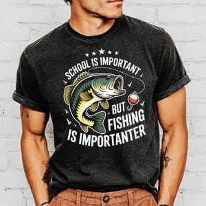 May include: Dark gray t-shirt with the text "SCHOOL IS IMPORTANT BUT FISHING IS IMPORTER". The shirt features a graphic of a fish with a fishing hook and bobber. The shirt has rolled-up sleeves.