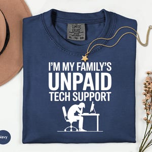 May include: Navy blue t-shirt with the text "I'M MY FAMILY'S UNPAID TECH SUPPORT" in white. The graphic shows a person at a desk with a computer. A gold necklace with a star pendant is on the shirt.