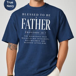 May include: True navy blue t-shirt with the text "BLESSED TO BE A FATHER" in white, along with a Bible verse. The shirt has a classic crew neck and short sleeves. The text "PROVERBS 20:7" is also visible.