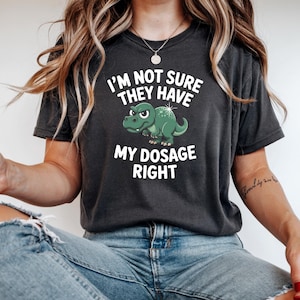 May include: Dark gray t-shirt with the text "I'M NOT SURE THEY HAVE MY DOSAGE RIGHT" in white. A green cartoon dinosaur is featured in the center. The shirt is a casual wear item.