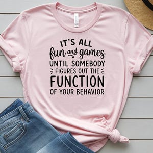 May include: A light pink t-shirt with the text "IT'S ALL fun and games UNTIL SOMEBODY FIGURES OUT THE FUNCTION OF YOUR BEHAVIOR" in black. The shirt is paired with blue jeans and a straw hat.