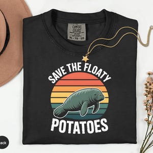 May include: Black t-shirt with a graphic of a manatee inside a sunset design. The text reads "Save the Floaty Potatoes." A gold necklace with a star pendant is draped over the shirt. The shirt is labeled XL.
