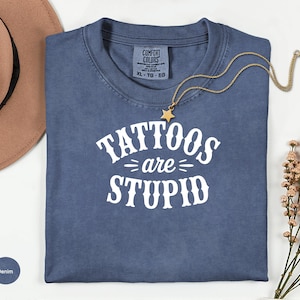 May include: Denim blue t-shirt with the text "Tattoos are Stupid" in white. A gold necklace with a star pendant is draped across the shirt. A brown hat and flowers are in the background.