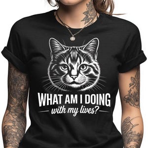May include: Black t-shirt featuring a detailed black and white illustration of a cat's face. Below the cat graphic, the text reads "WHAT AM I DOING with my lives?" in a bold, white font. The shirt has short sleeves.