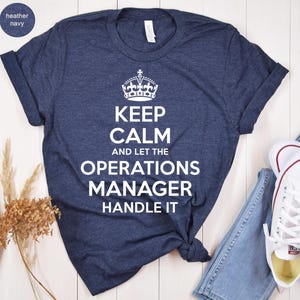 May include: Heather navy blue t-shirt with the text "KEEP CALM AND LET THE OPERATIONS MANAGER HANDLE IT" in white. The shirt features a crown graphic above the text. The shirt is styled with rolled sleeves.