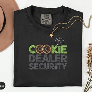 May include: Black crew neck t-shirt with the text "COOKIE DEALER SECURITY" in green, brown, and gray. The design includes donut graphics and a siren. A gold necklace with a star pendant is draped on the shirt.