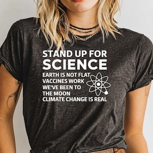 May include: Dark gray t-shirt with the text "STAND UP FOR SCIENCE" and the statements "EARTH IS NOT FLAT, VACCINES WORK, WE'VE BEEN TO THE MOON, CLIMATE CHANGE IS REAL". A white atom symbol is also present.