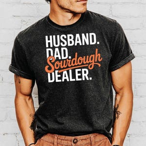 May include: Dark gray t-shirt with the words "HUSBAND. DAD. Sourdough DEALER." in white and orange lettering. The shirt has rolled-up sleeves and is worn by a person.