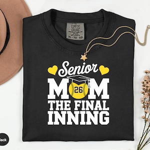 May include: Black t-shirt with the text "Senior Mom" and a baseball graphic. The baseball has the number "26" on it. The shirt also has the words "The Final Inning" in white. A gold necklace with a star pendant is on the shirt.