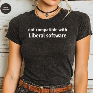 May include: Black heather t-shirt with the text "not compatible with Liberal software" in white. The shirt has a crew neck and short sleeves. The model is wearing a black choker necklace and a brown belt.