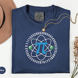 May include: Navy blue t-shirt with a white and blue Pi symbol design. The design includes the Pi symbol, numbers, and atom-like shapes. A gold necklace with a star pendant is on the shirt. The shirt is a Comfort Colors XL.