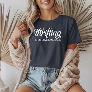 May include: A navy blue t-shirt with the white text "thrifting IS MY LOVE LANGUAGE". The shirt is short-sleeved and has a crew neck. The model is wearing denim shorts and a beige cardigan.