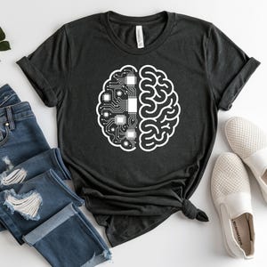 May include: Dark gray t-shirt featuring a white brain graphic. The graphic is split in half, with one side depicting a circuit board and the other side a brain. The shirt is paired with blue jeans and white slip-on shoes.