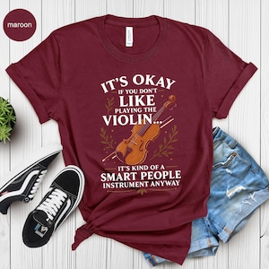 May include: Maroon t-shirt with the text "It's Okay If You Don't Like Playing the Violin... It's Kind of a Smart People Instrument Anyway" and a violin graphic. The shirt is paired with denim shorts and black sneakers.