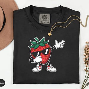 May include: Black crew neck t-shirt featuring a cartoon strawberry wearing sunglasses and doing a dab pose. The strawberry has white gloves and shoes. A gold necklace with a star pendant is also visible.