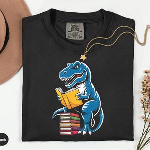 May include: Black t-shirt featuring a blue cartoon dinosaur reading a yellow book. The dinosaur is sitting on a stack of colorful books. A gold necklace with a star pendant is draped over the shirt.