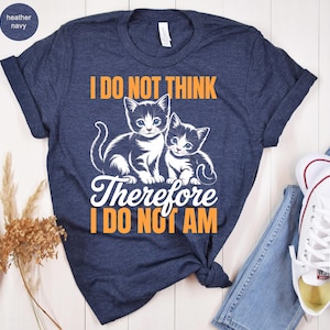 May include: Heather navy t-shirt featuring two kittens and the text "I DO NOT THINK Therefore I DO NOT AM" in orange and white. The shirt is paired with blue jeans and white sneakers.